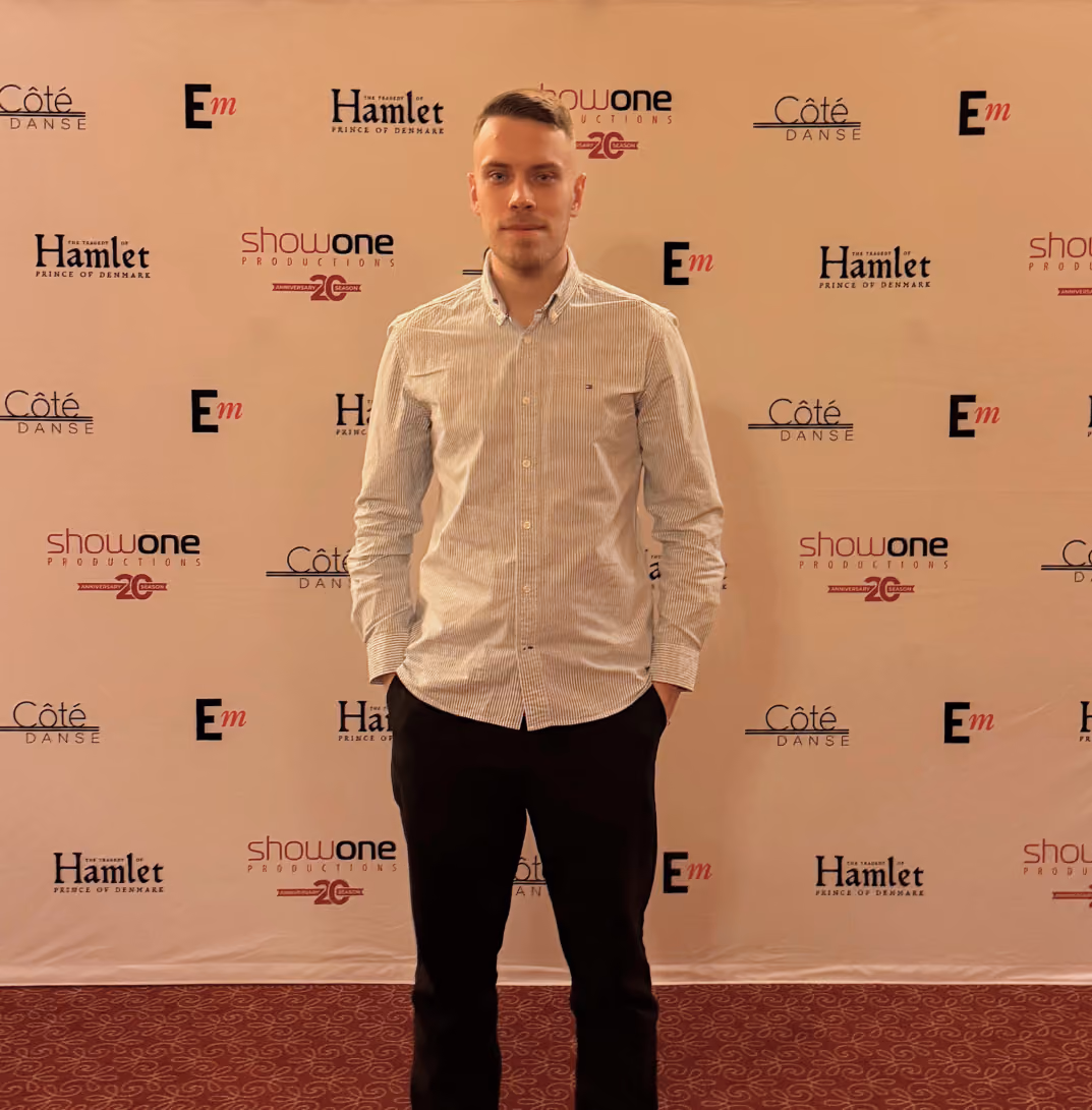 Yaroslav stands on a red patterned carpet in front of a beige backdrop with logos for Côté Danse, Hamlet Prince of Denmark, show one productions, and Em repeated in a grid pattern.