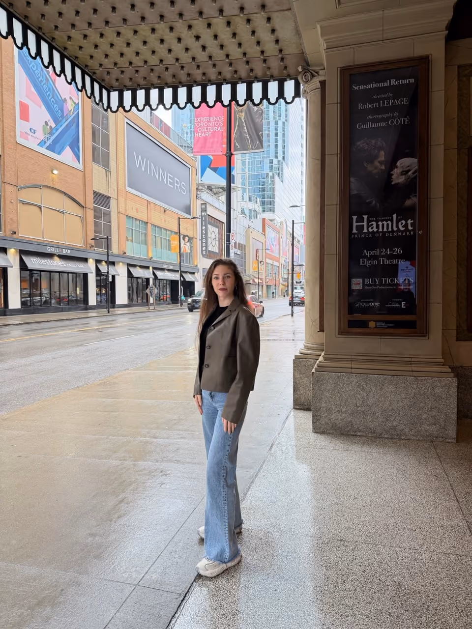Kateryna stands on a city sidewalk under a marquee with light bulbs in front of a theater advertising a production of Hamlet at the Elgin Theatre.