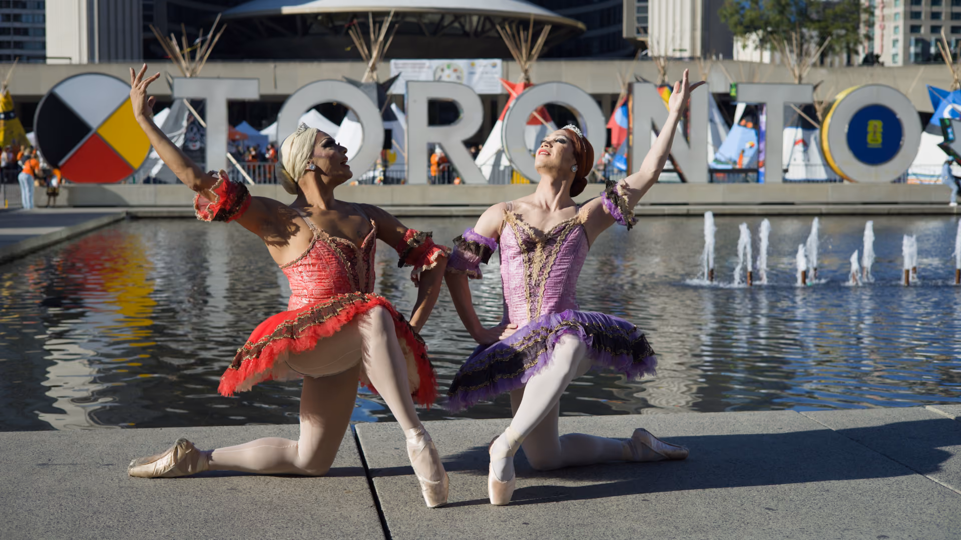 Two ballet dancers kneeling in front of the rainbow-colored Toronto sign near a fountain, one wearing a red tutu and the other a purple tutu, both posing with arms raised.