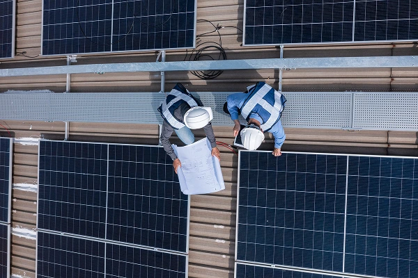 Técnicos con cascos y chalecos revisando planos mientras instalan paneles solares en un techo.