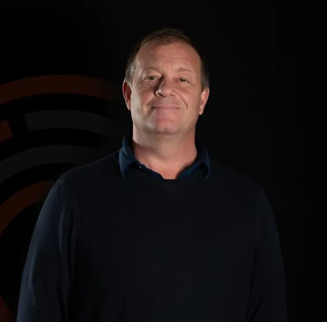 Smiling middle-aged man wearing a dark sweater over a blue collared shirt against a black background.