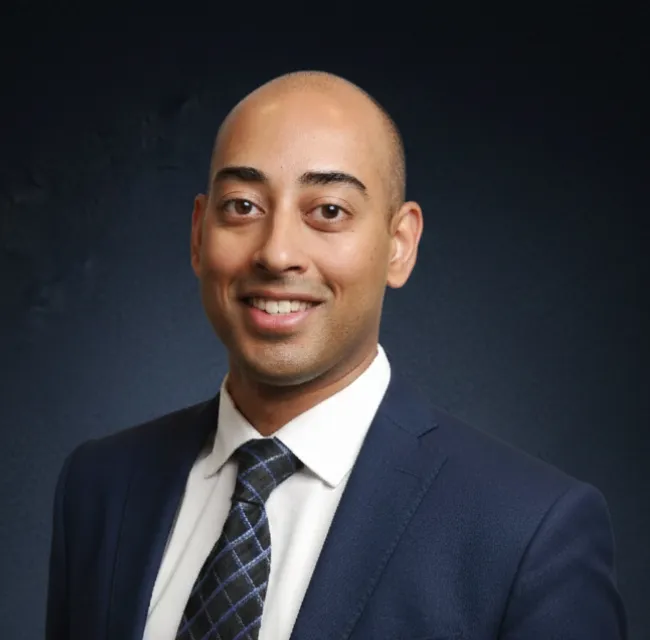 Smiling bald man wearing a dark blue suit, white shirt, and patterned tie against a dark background.