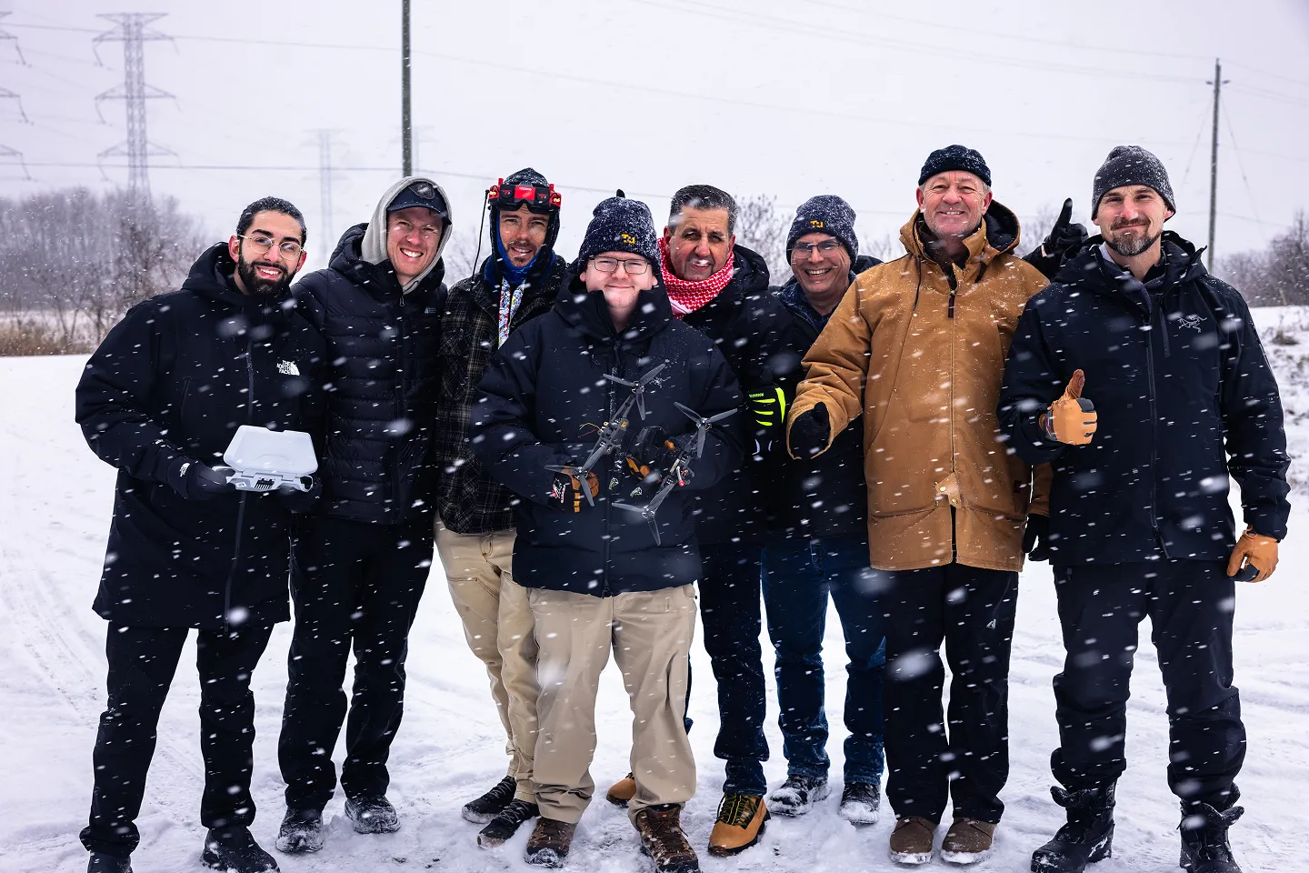 Group of eight men standing in snow during snowfall, wearing winter clothing, with one holding a drone and another holding a remote controller.