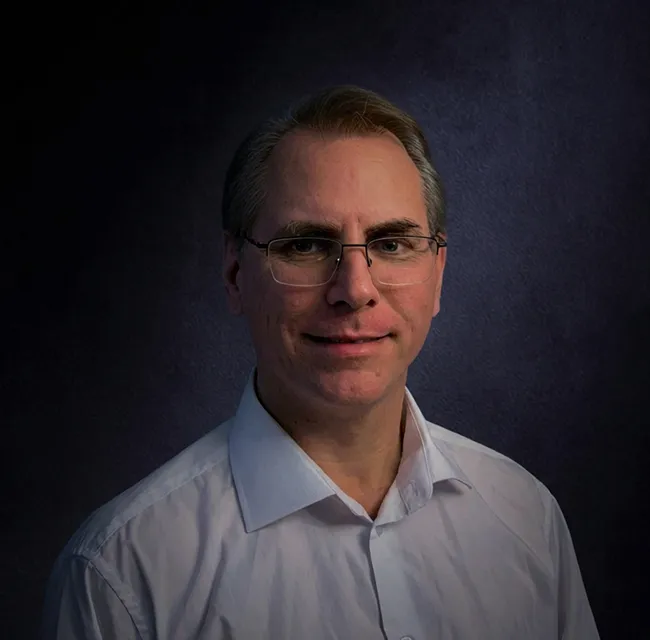 Portrait of a middle-aged man wearing glasses and a white collared shirt against a dark background.