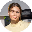 Portrait of a woman with long brown hair in a light yellow sweater, smiling slightly in an office setting.