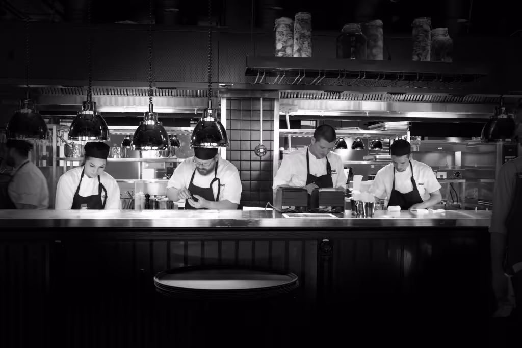 Fyra kockar iklädda vita skjortor och svarta förkläden jobbar i ett restaurangkök med hängande lampor ovanför arbetsytan.
