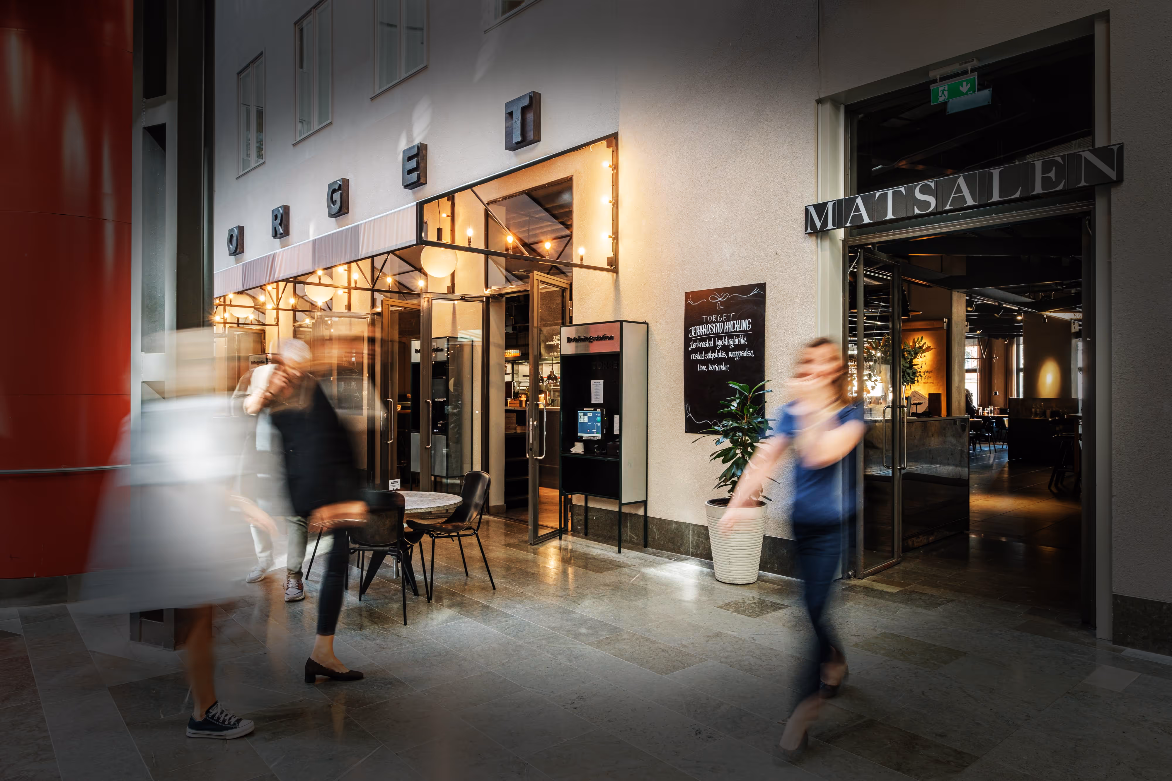 Personer i rörelse framför ingången till en restaurang med skyltar som lyder 'TORGET' och 'MATSALEN'.