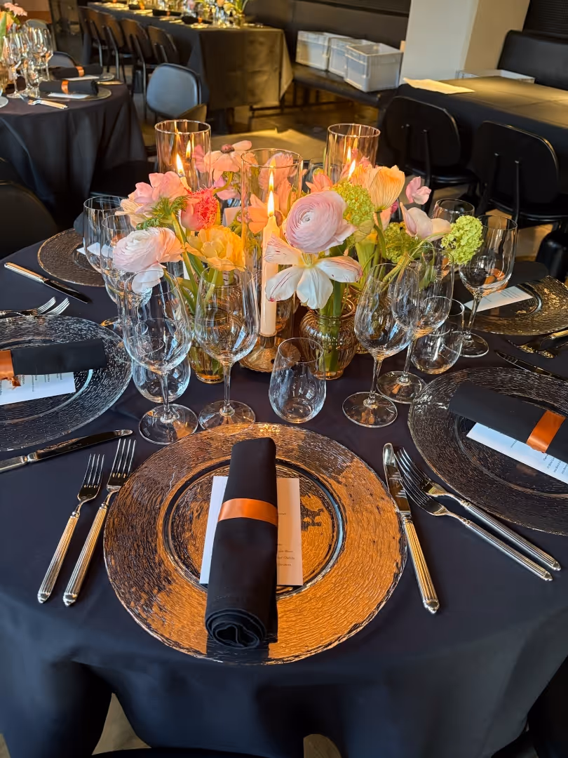 Elegant dining table with black tablecloth, glass charger plates, black napkins with copper rings, multiple wine glasses, and a floral centerpiece with lit candles.
