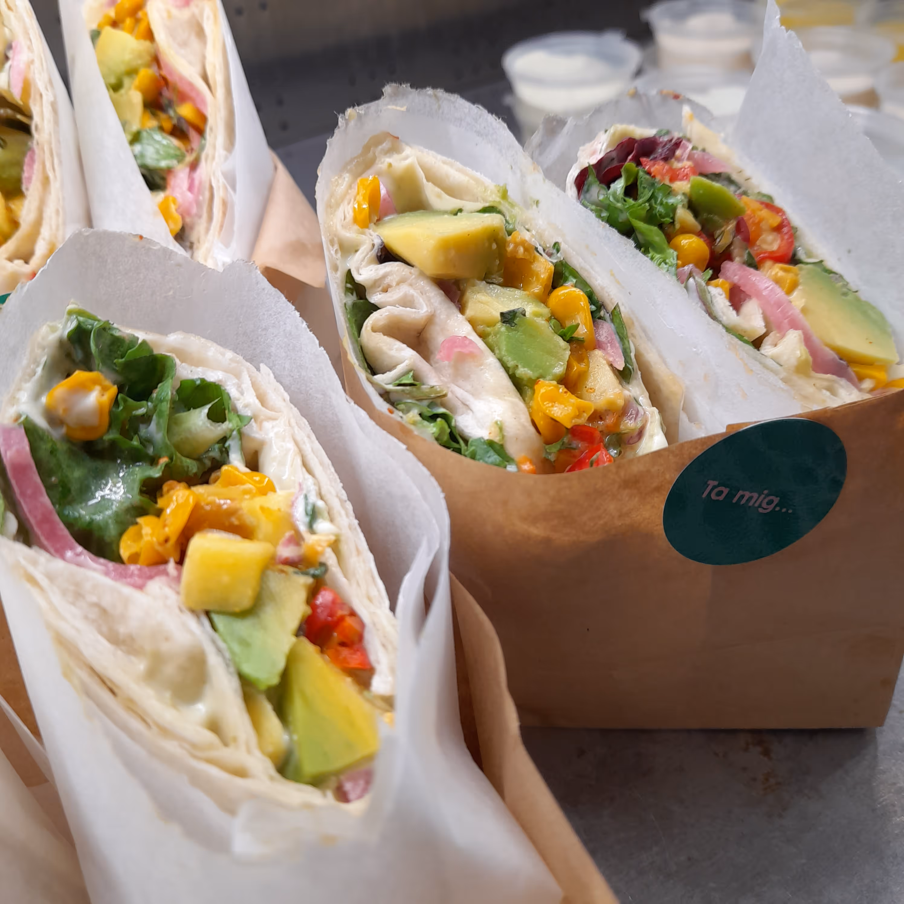 Flera wraps fyllda med avokado, majs, sallad och rödlök inslagna i papper och placerade i bruna pappershållare.