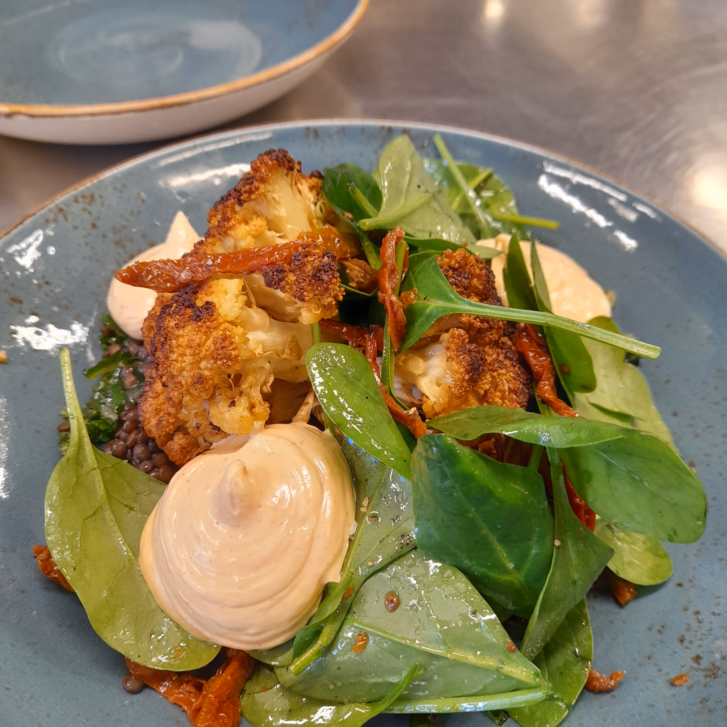 Rostad blomkål och spenat med krämig sås och soltorkade tomater på blå tallrik.