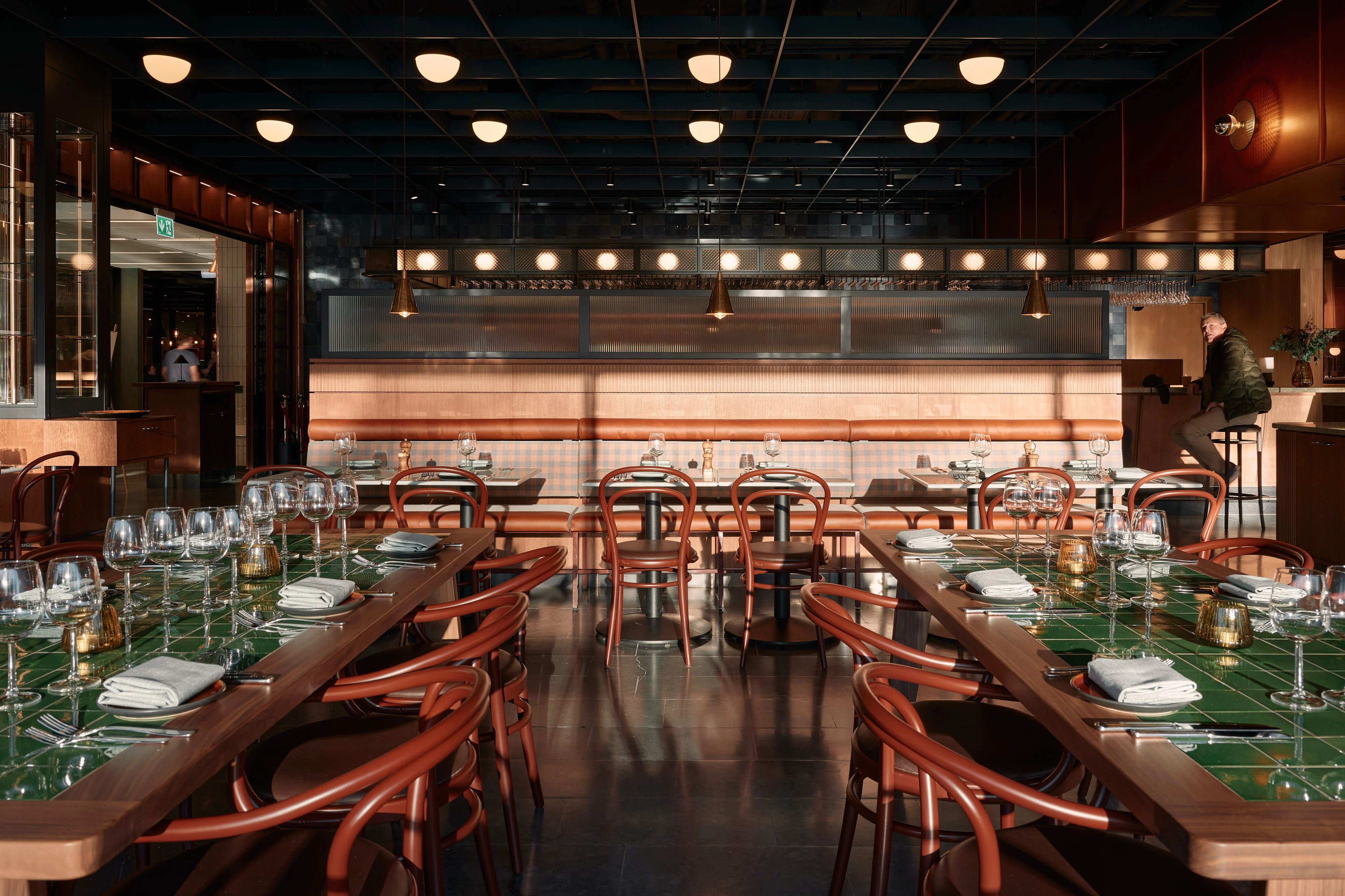 Ljust och modernt restauranginteriör med gröna kaklade bord dukade med vinglas, bruna stolar och en man som sitter vid en bardisk.