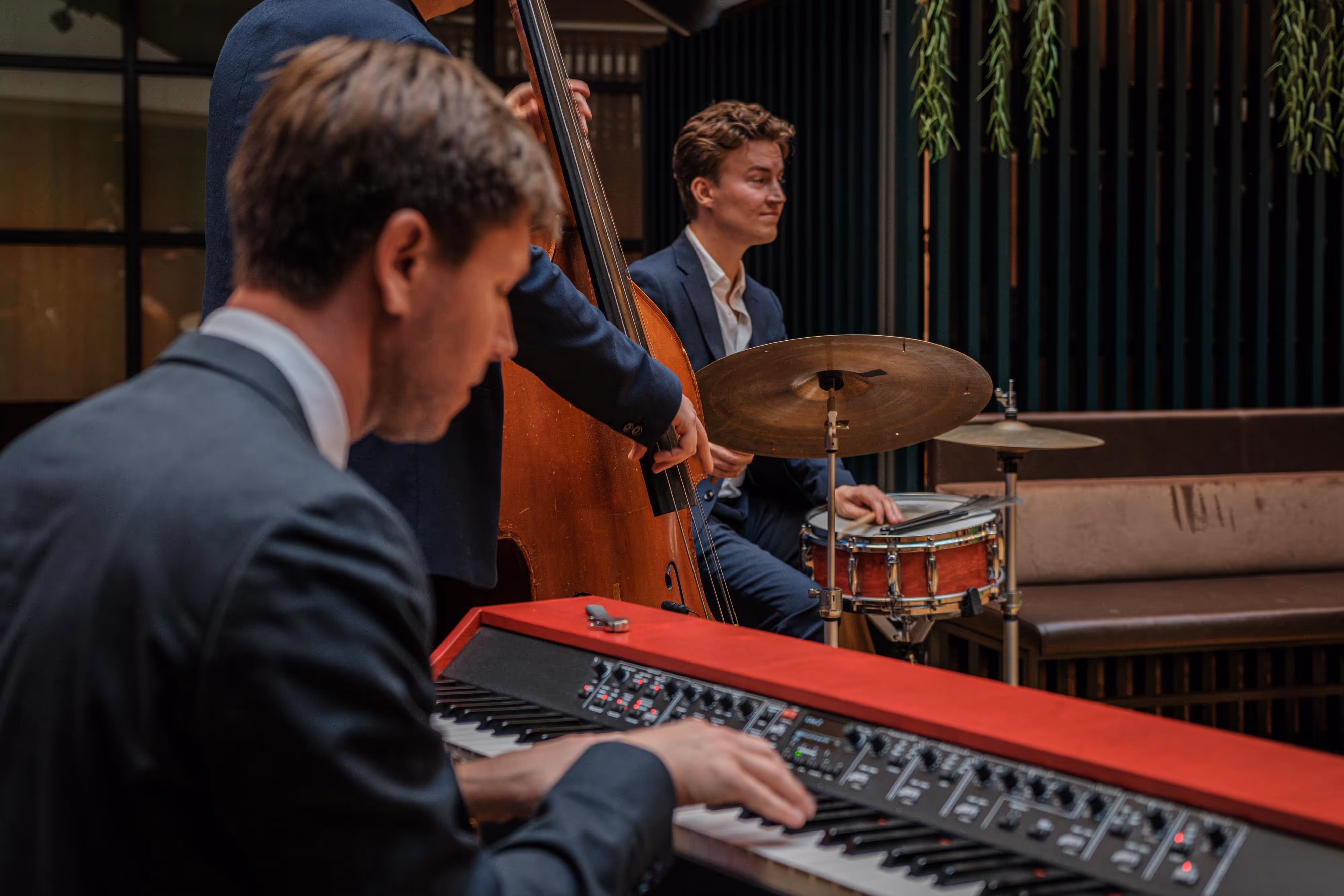 Tre män i kostym spelar jazzmusik med keyboard, kontrabas och trummor inomhus.