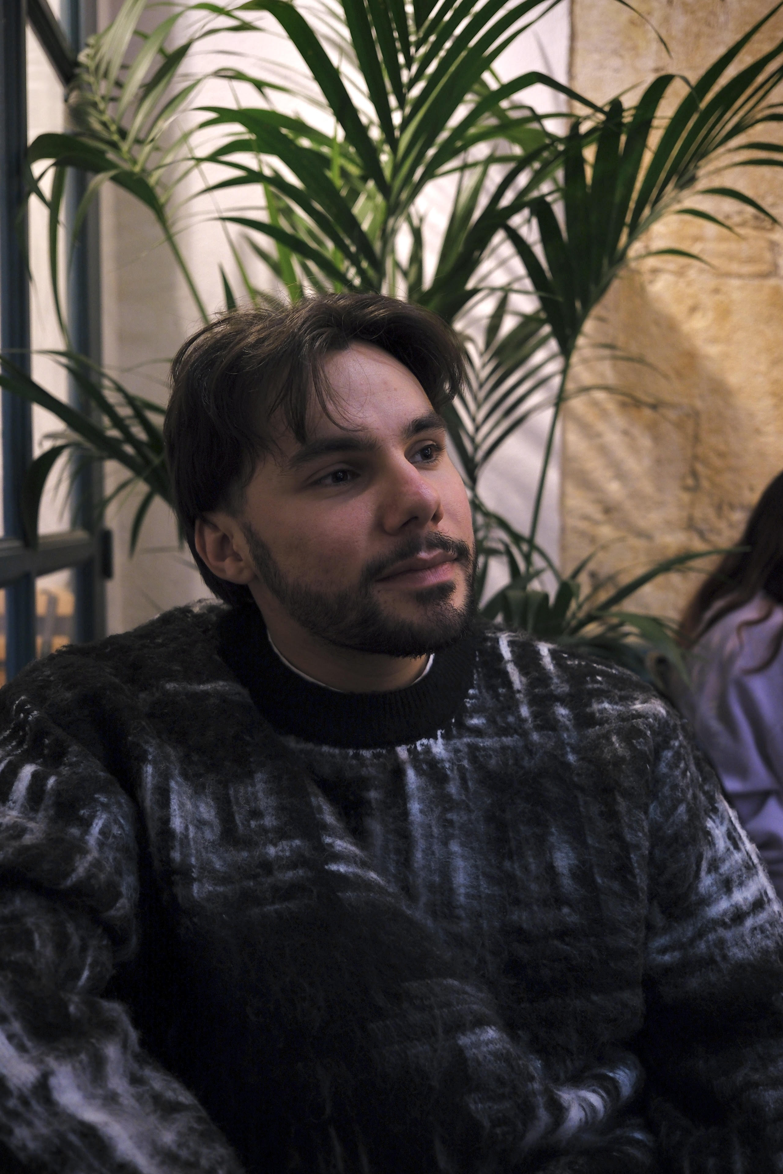 Jeune homme barbu portant un pull noir et gris, assis à l'intérieur devant des grandes plantes vertes, regardant vers la droite.