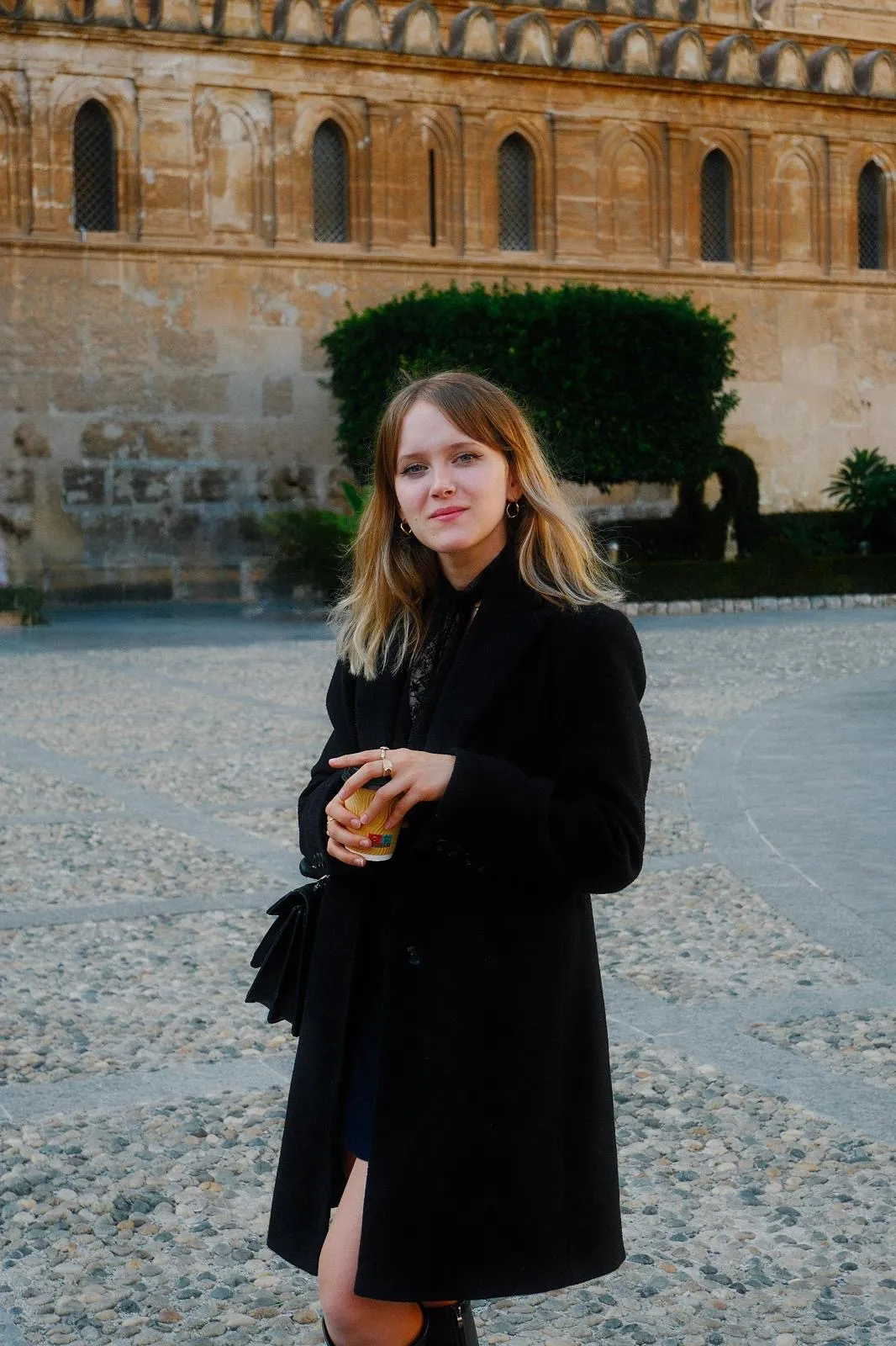 Jeune femme aux cheveux blonds portant un manteau noir, tenant une tasse en carton dans un cadre urbain avec un mur ancien en arrière-plan.