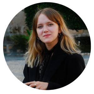 Portrait d'une jeune femme aux cheveux blonds, portant un manteau noir, avec un fond flou de verdure et mur.