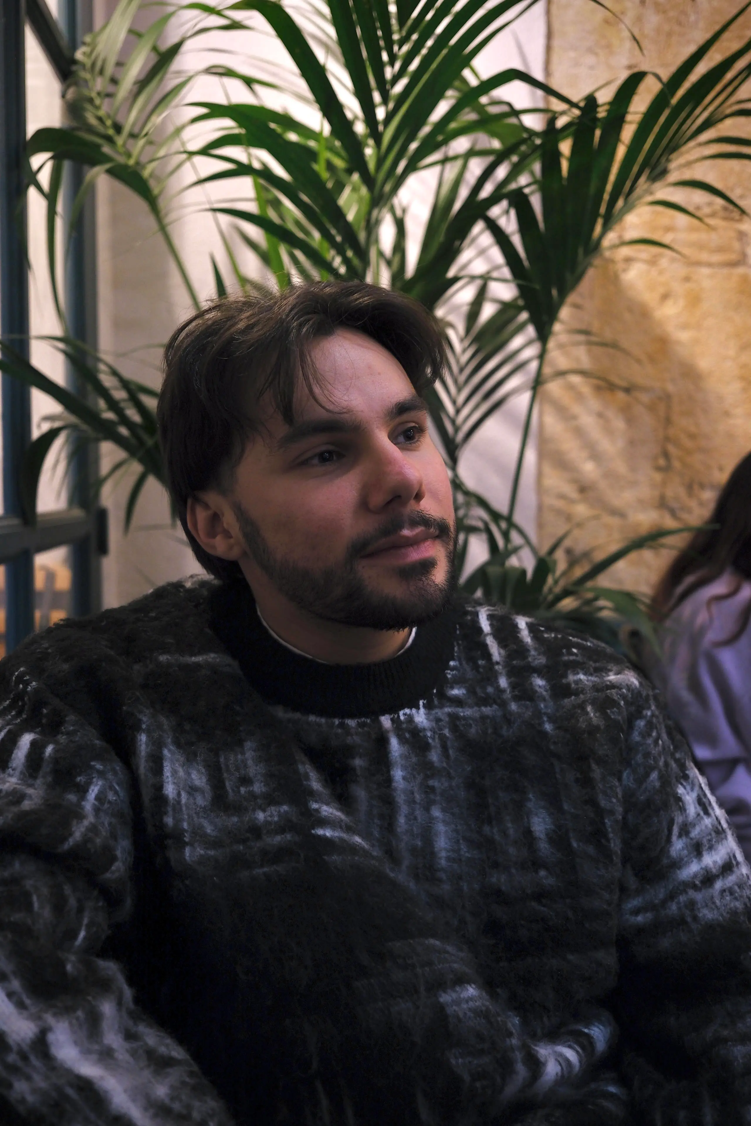 Jeune homme barbu en pull à motifs sombres assis devant des plantes vertes en intérieur.