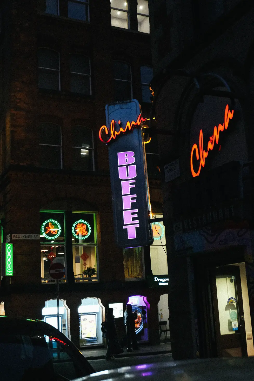 Enseigne lumineuse de nuit indiquant un buffet chinois sur un bâtiment urbain avec silhouettes de passants.