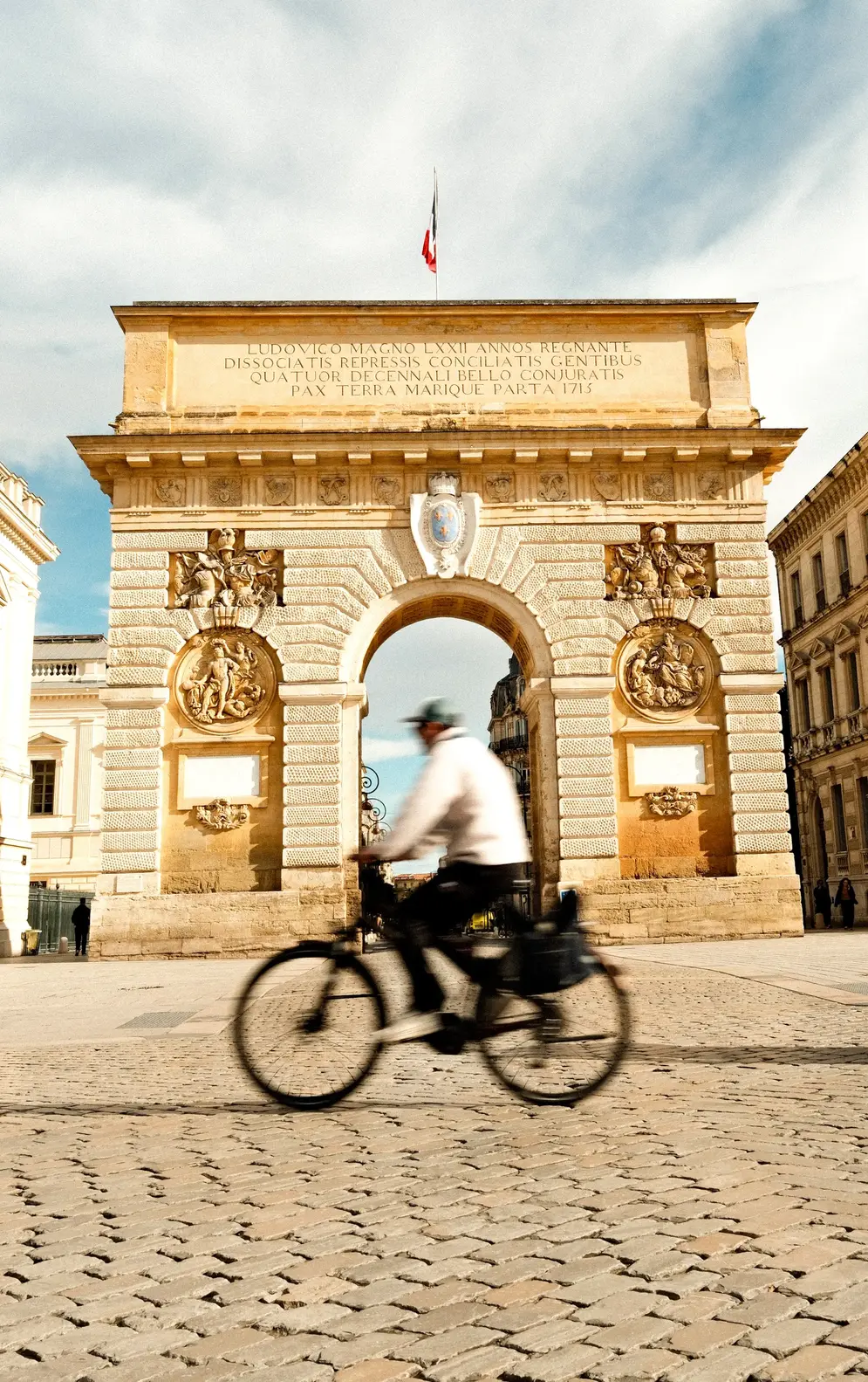 Arc de triomphe en pierre avec sculptures et drapeau français au sommet, cycliste flou passant devant sur pavés.