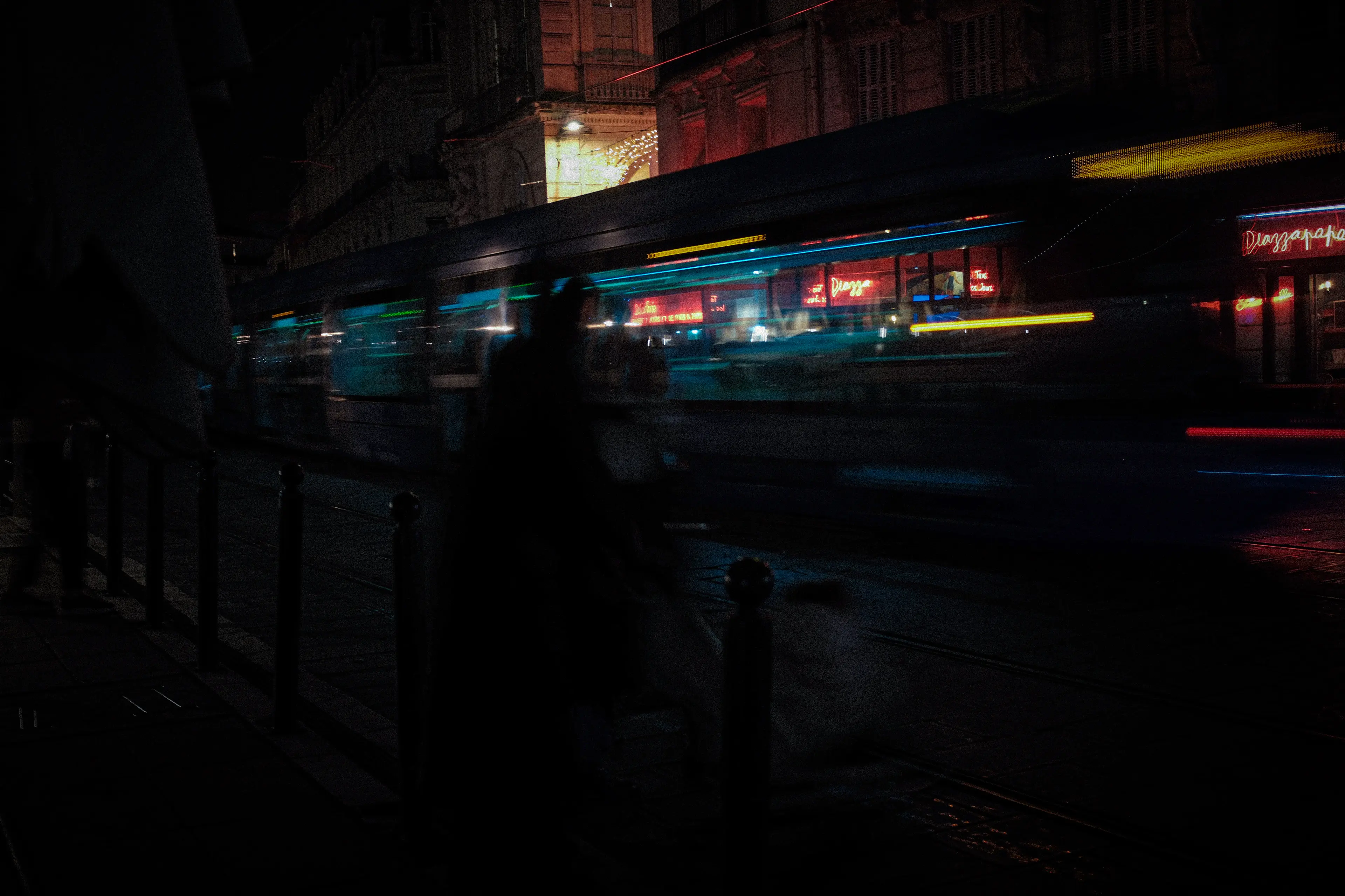 Tram en mouvement flou la nuit devant un bâtiment éclairé avec des enseignes néon rouges.