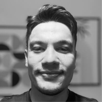 Yakından çekilmiş siyah-beyaz bir erkek yüzü, hafif gülümseyen ve arkada soyut desenli resimler bulunan.