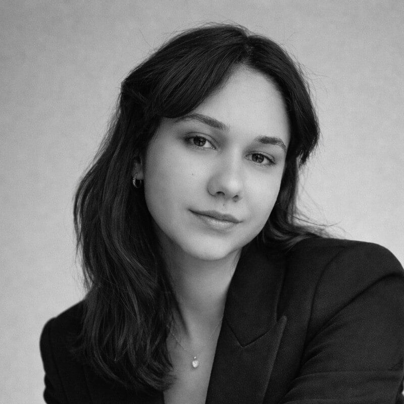 Uzun koyu saçlı ve siyah blazer ceket giymiş, hafif gülümseyen genç kadın portresi.