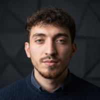 Kısa kıvırcık saçlı, sakallı genç adam siyah kazakla, koyu geometrik desenli arka planda.