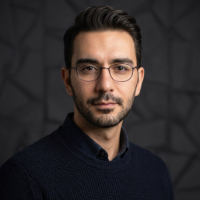 Gözlüklü ve koyu kazak giymiş, karanlık desenli arka plan önünde ciddi ifadeli erkek portresi.