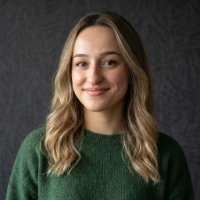 Koyu gri arka plan önünde yeşil kazak giymiş, hafif dalgalı saçlı, gülen genç kadın.