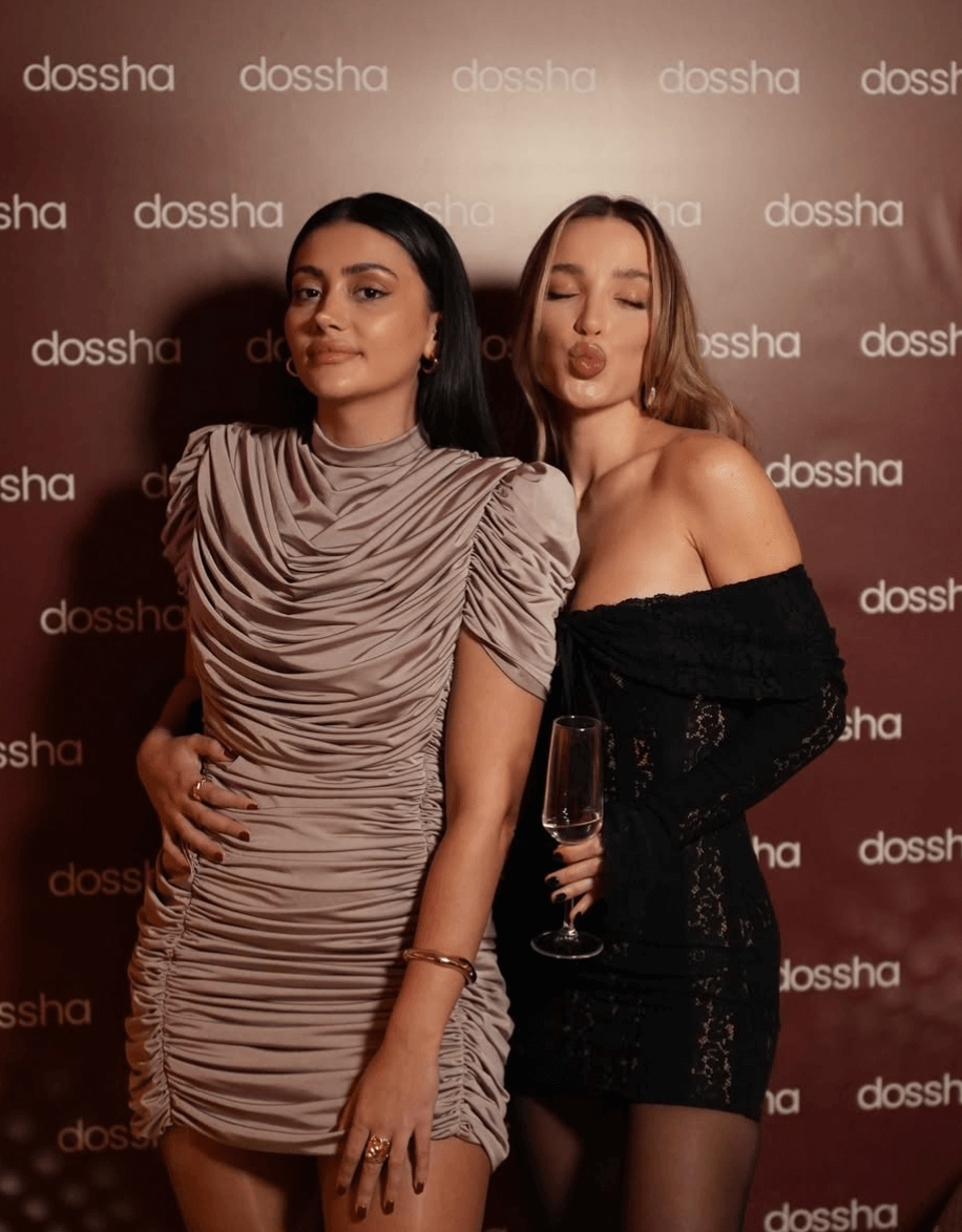 İki kadın dosscha yazılı kahverengi arka önünde, biri bej rütüşlü kısa elbise giymiş, diğeri siyah dantel omuzdan açık elbise içinde kadeh tutuyor ve öpücük pozu veriyor.