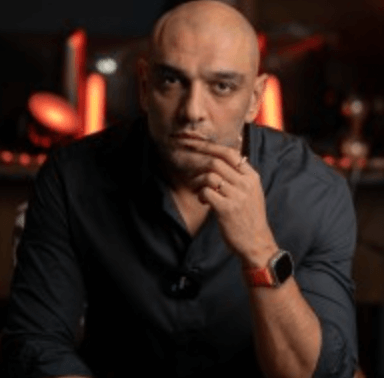 Düşünceli bir ifadeyle çenesine parmağını dayamış, koyu renk gömlek giymiş kel bir adam.