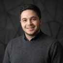 Gri kazak giymiş, koyu saçlı ve sakallı genç erkek, gri geometrik desenli arka planda gülümseyerek poz veriyor.