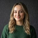 Gri arka plan önünde yeşil kazak giymiş, açık kahverengi dalgalı saçlı ve hafif gülümseyen genç kadın portresi.