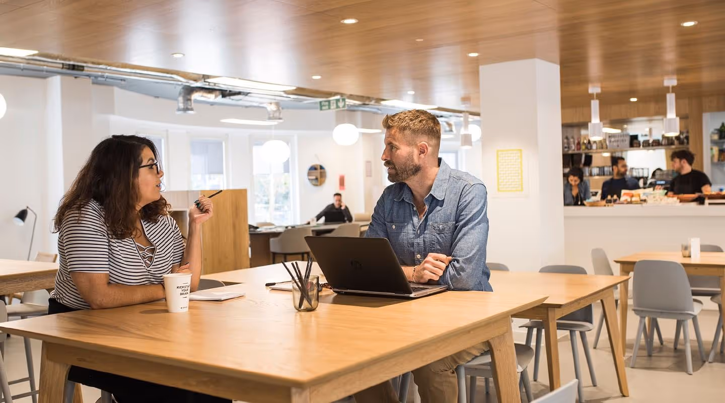 Two people having a discussion at a wooden table in a bright modern coworking space with a laptop and coffee cup.