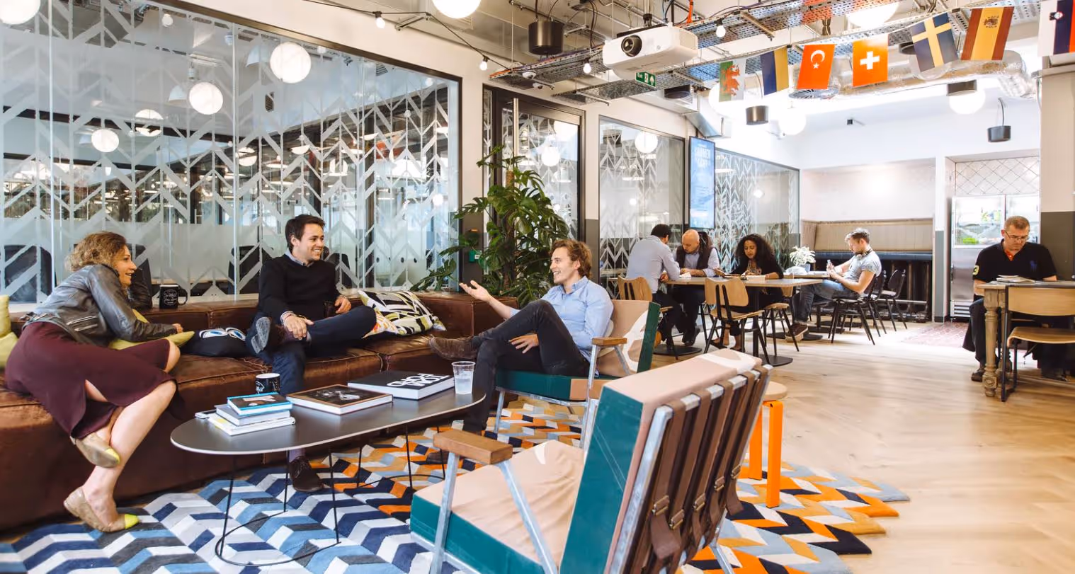 Modern coworking space with people sitting on sofas and chairs chatting and others working at tables under hanging international flags and ceiling lights.