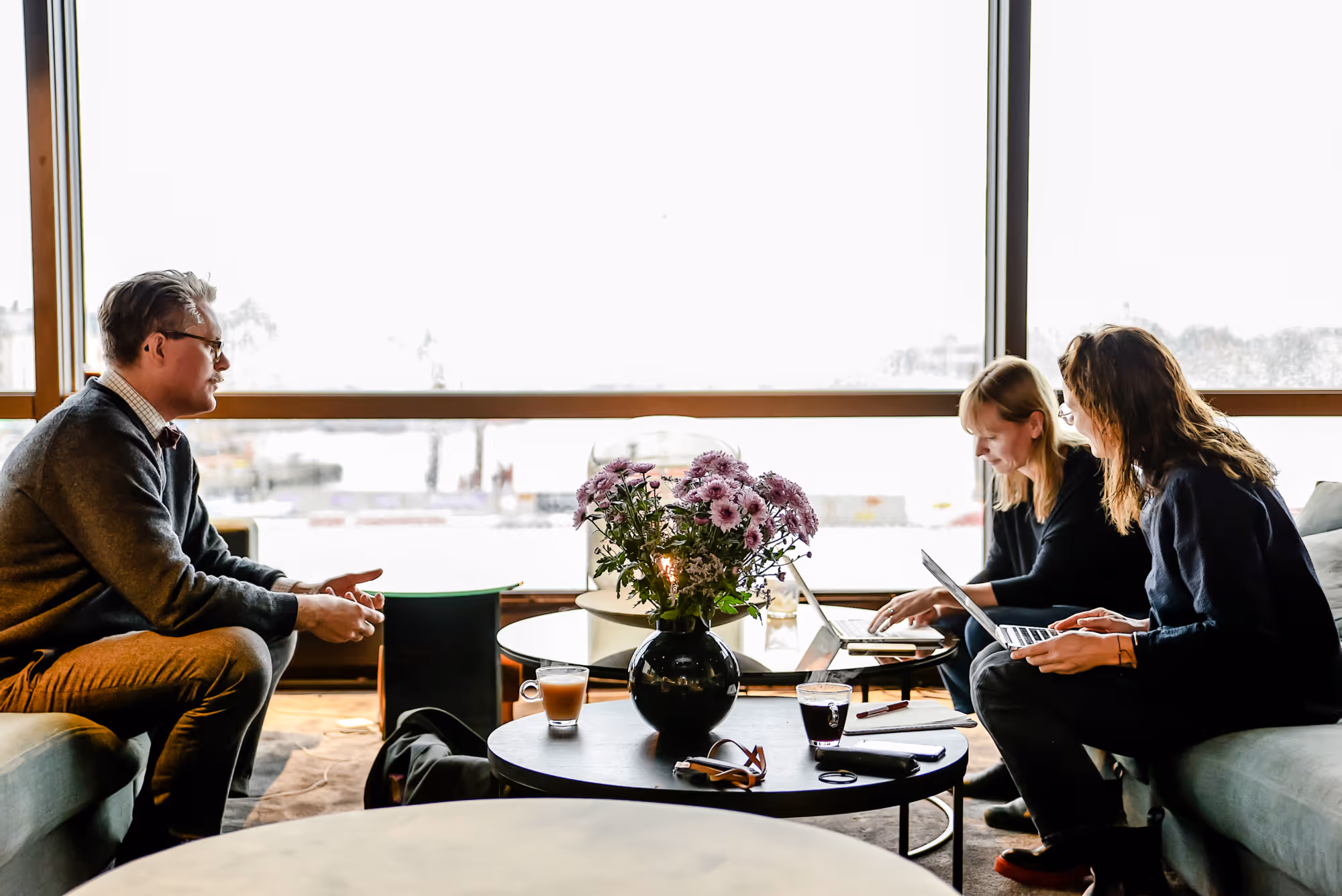 Three people sitting in a modern lounge with laptops and beverages on tables, discussing with a large window overlooking a bright outdoor scene in the background.