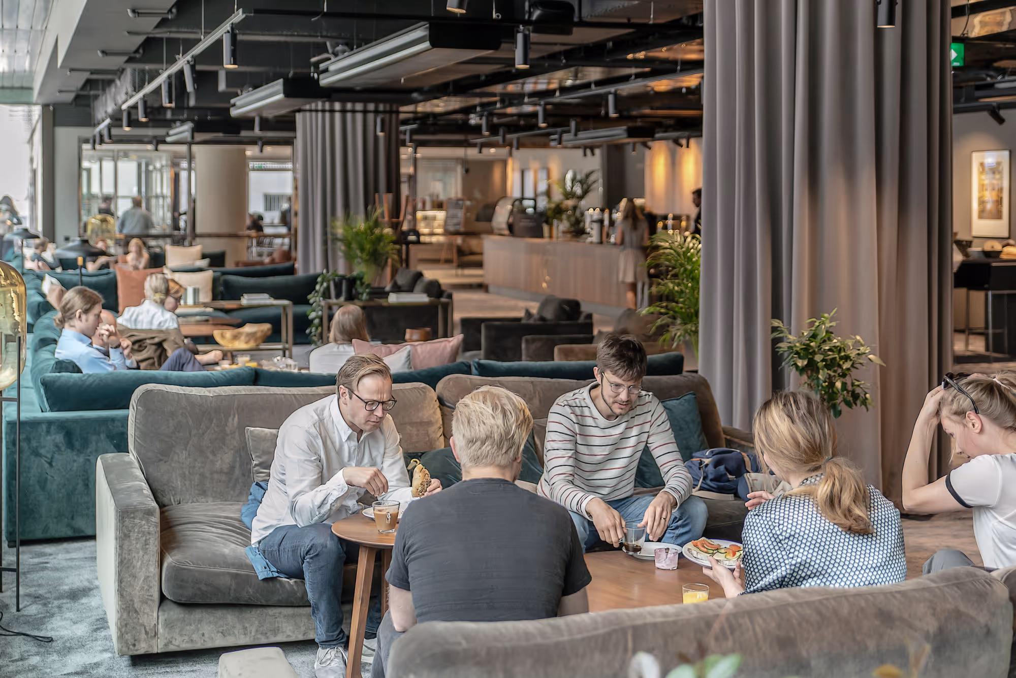 Group of five people sitting on sofas around a low table with drinks and snacks in a modern, spacious lounge area.