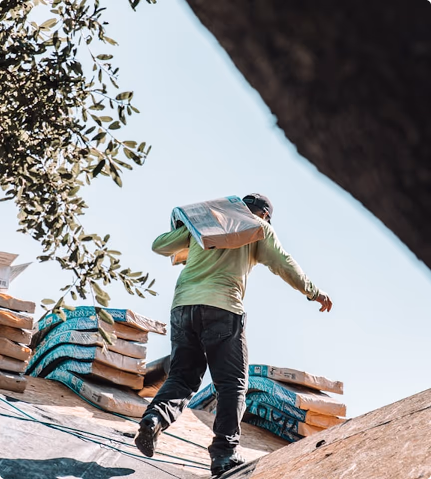 Worker carrying a bag of materials on the shoulder while walking on a rooftop under clear sky.