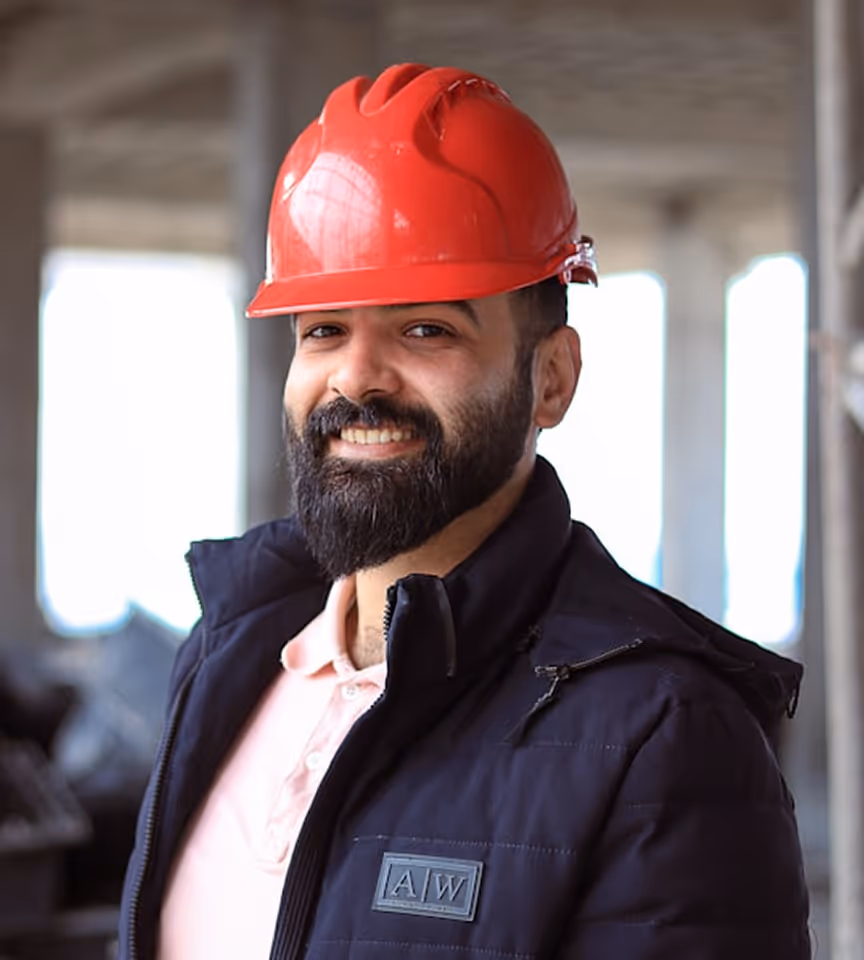 Smiling construction worker wearing a red hard hat and dark jacket standing inside a building under construction.