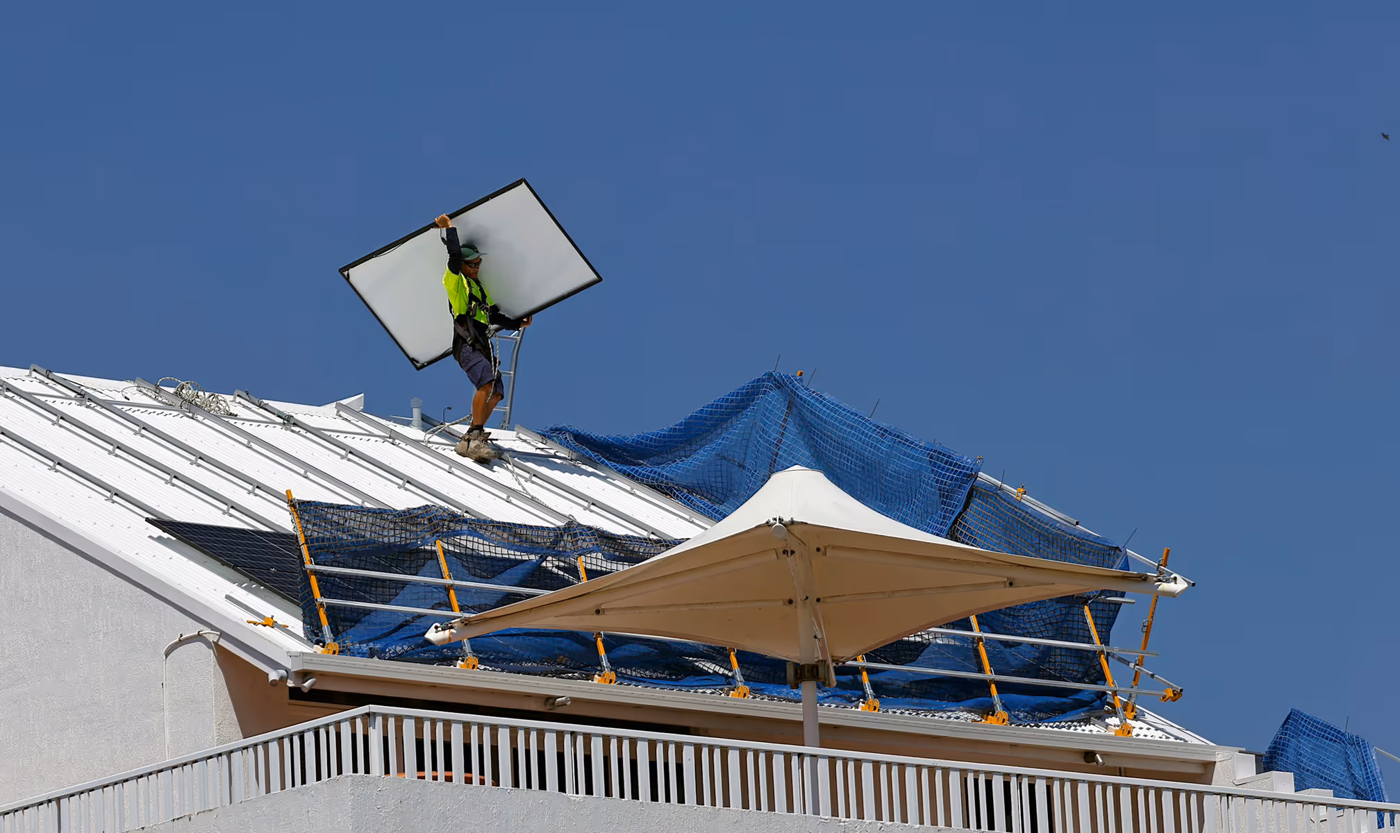 Worker in safety gear holding a solar panel while standing on a white metal roof under clear blue sky.