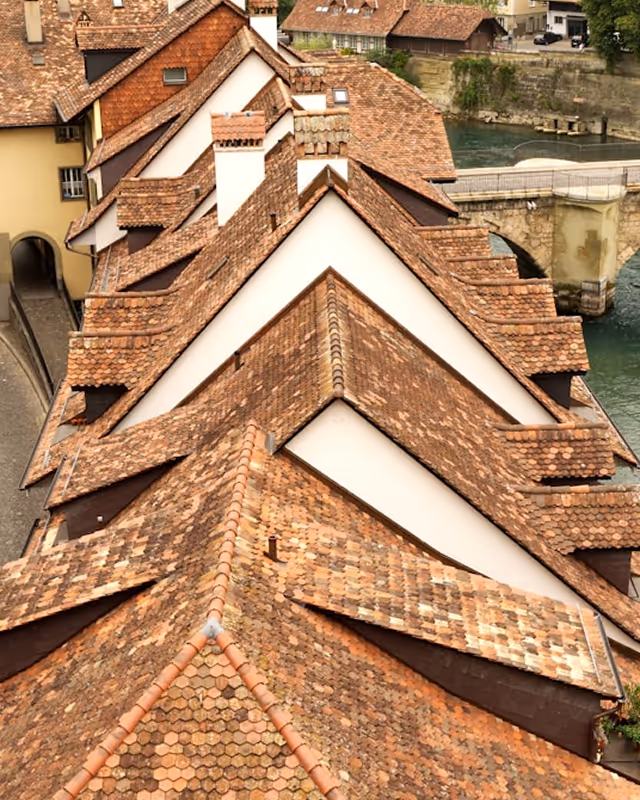 Top view of a building with multiple overlapping brown tiled roofs next to a river and stone bridge.