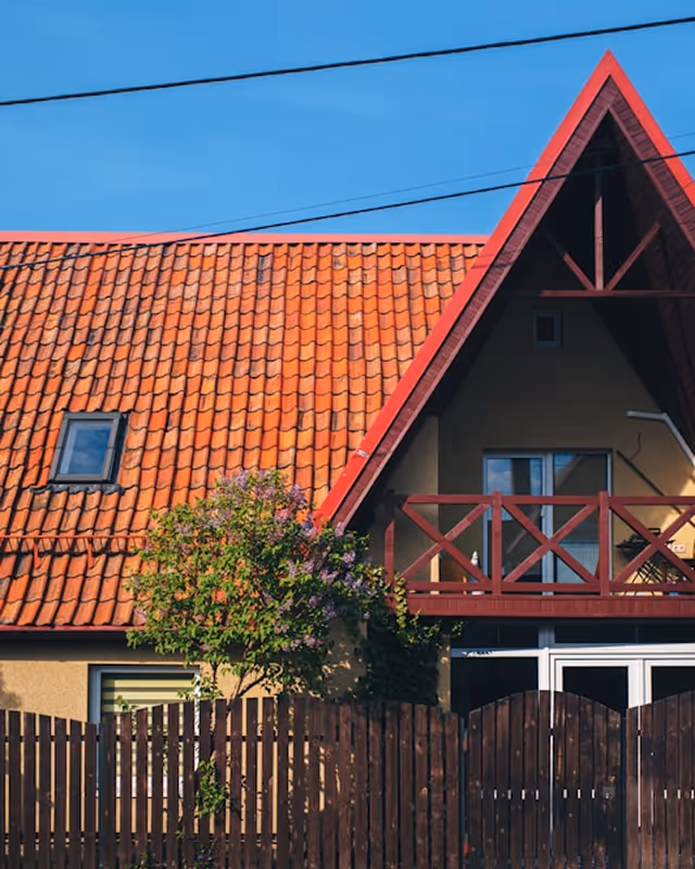 House with orange tiled roof, an A-frame balcony with red trim, and a wooden fence with a small tree in front under a clear blue sky.