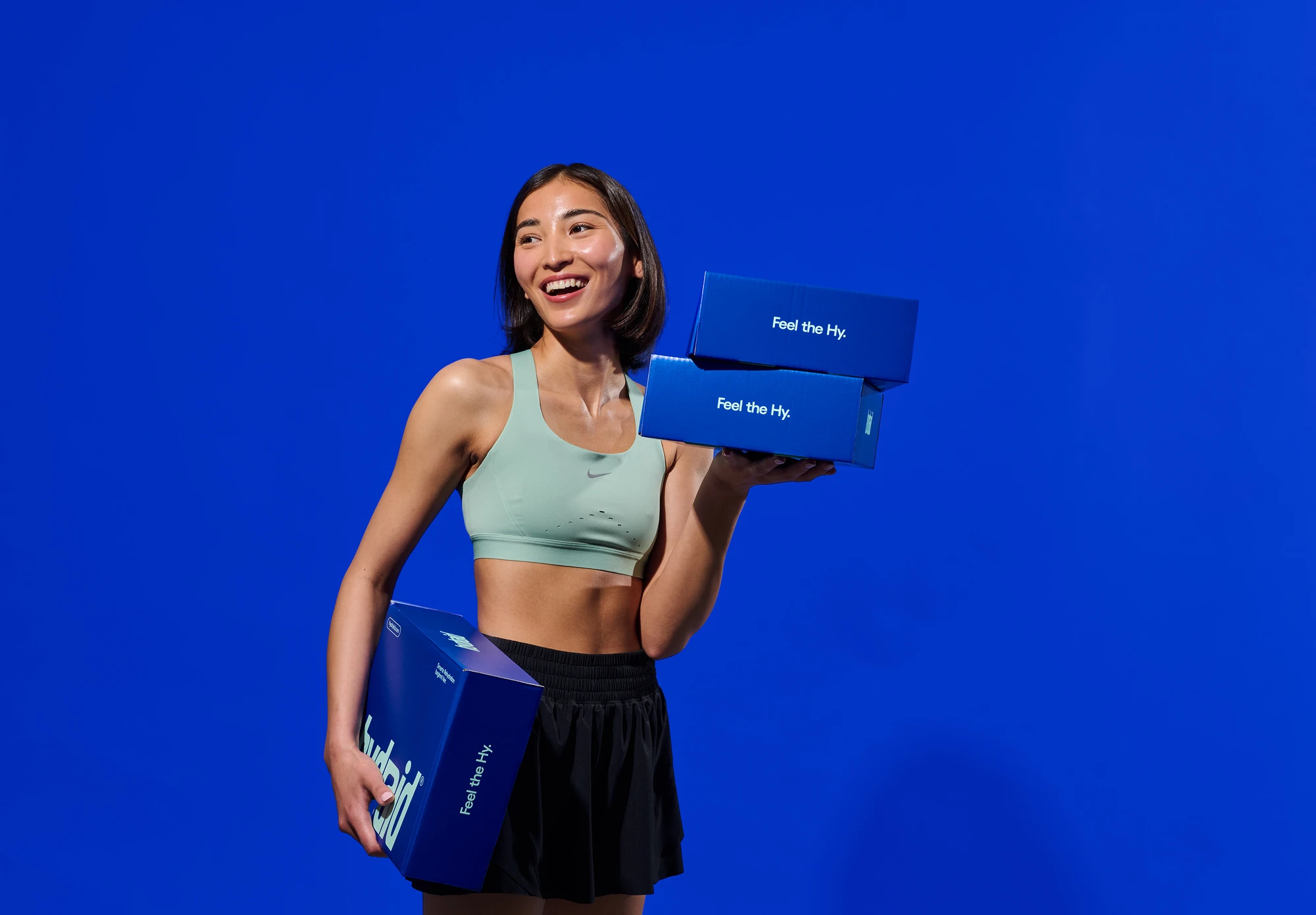 Werbefotografie für Hydraid: lächelnde Sportlerin hält Produktverpackungen vor blauem Studio-Hintergrund Advertising photography for Hydraid: smiling athlete holding product boxes against a blue studio backdrop