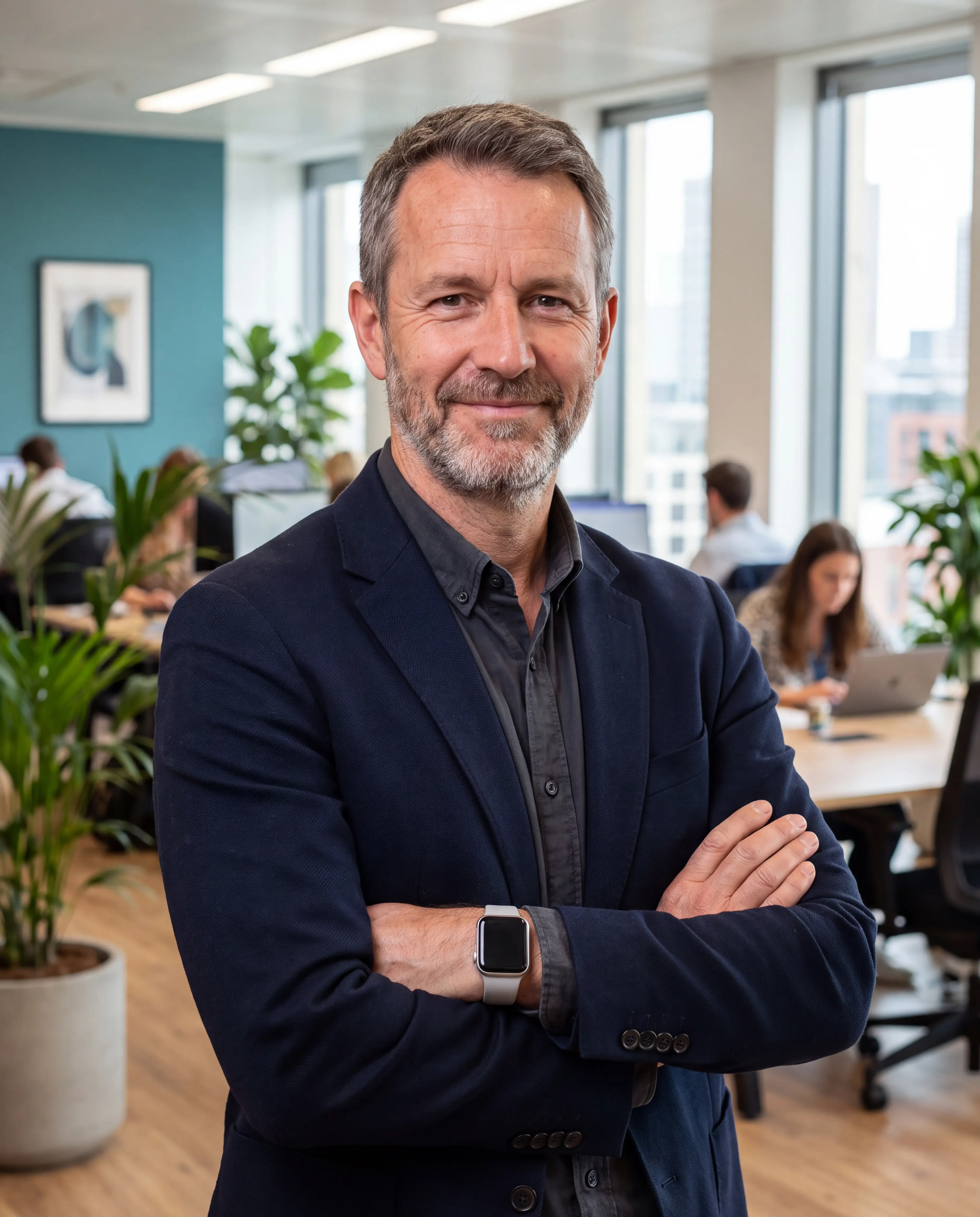 Confident man in a dark green blazer standing with arms crossed in a modern office with people working in the background.