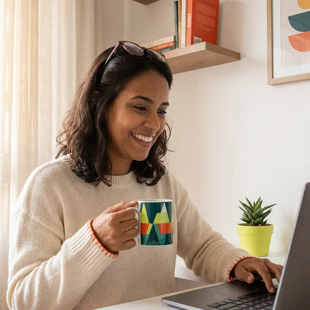 Smiling woman wearing a light sweater and sunglasses on her head, holding a colorful mug and working on a laptop at a desk with a small potted plant.