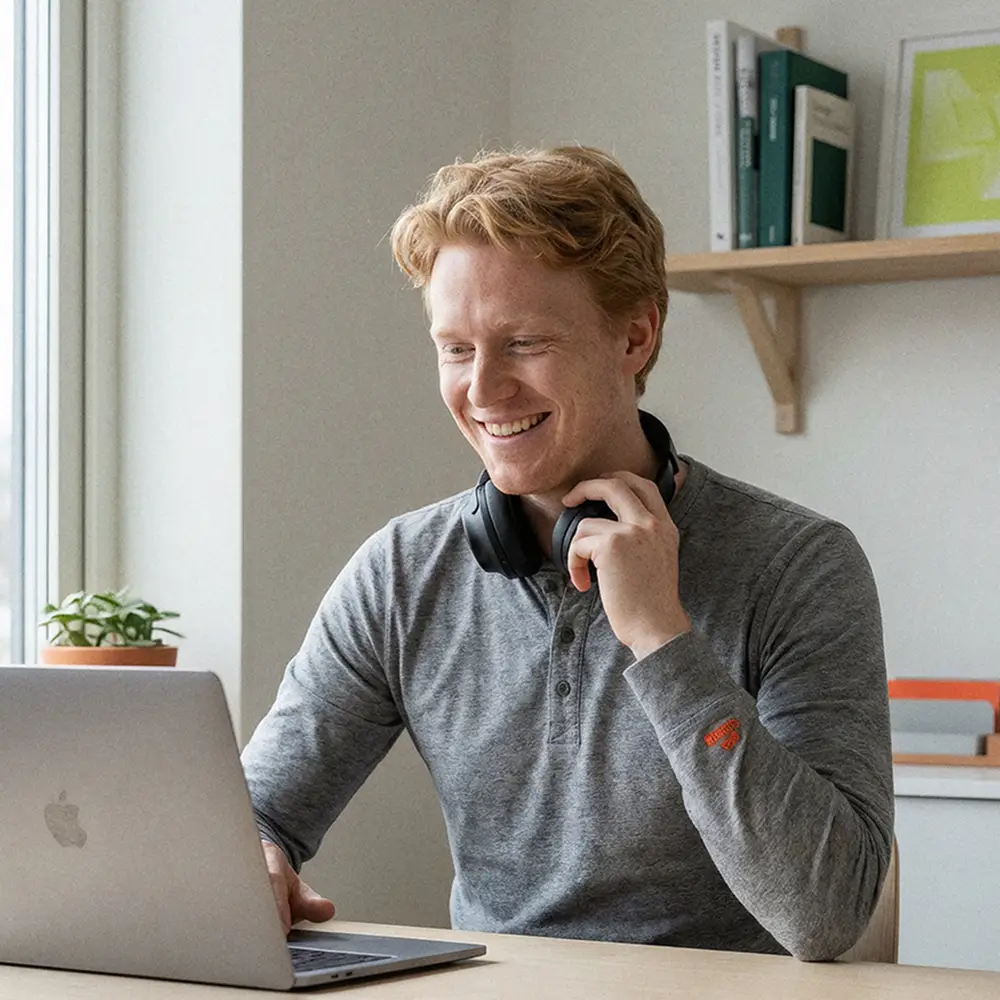 Smiling man in grey shirt using a laptop with headphones around his neck at a wooden desk near a window.