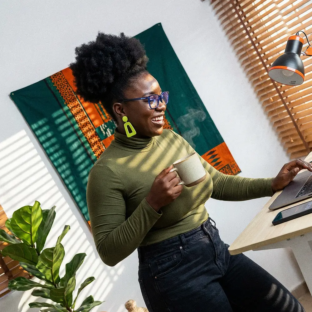 Woman with a green turtleneck holding a steaming mug, smiling at a laptop on a desk in a sunlit room with a decorative cloth on the wall.