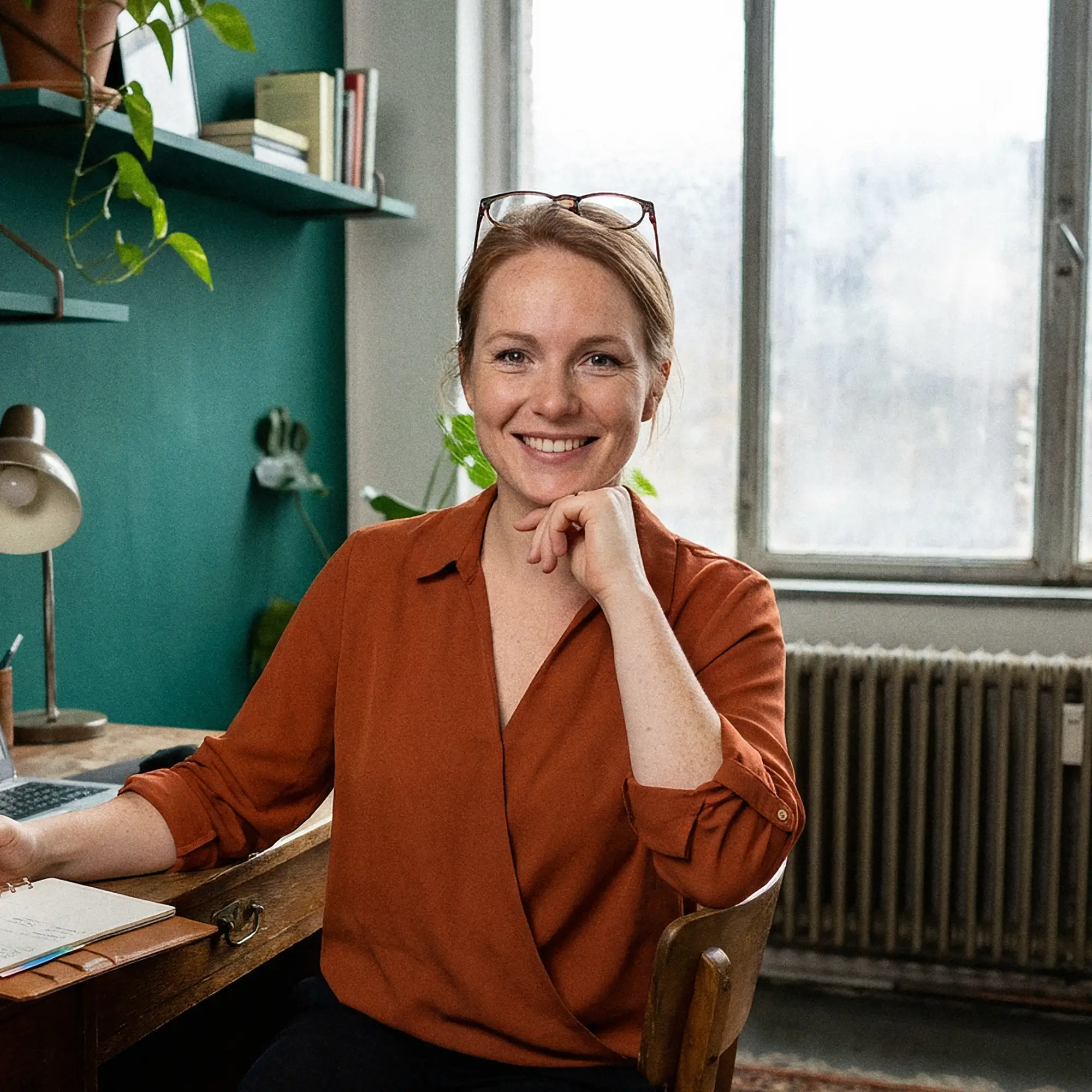 Smiling woman with light skin and red hair seated at a wooden desk with a notebook, wearing an orange blouse and glasses on her head in a home office.