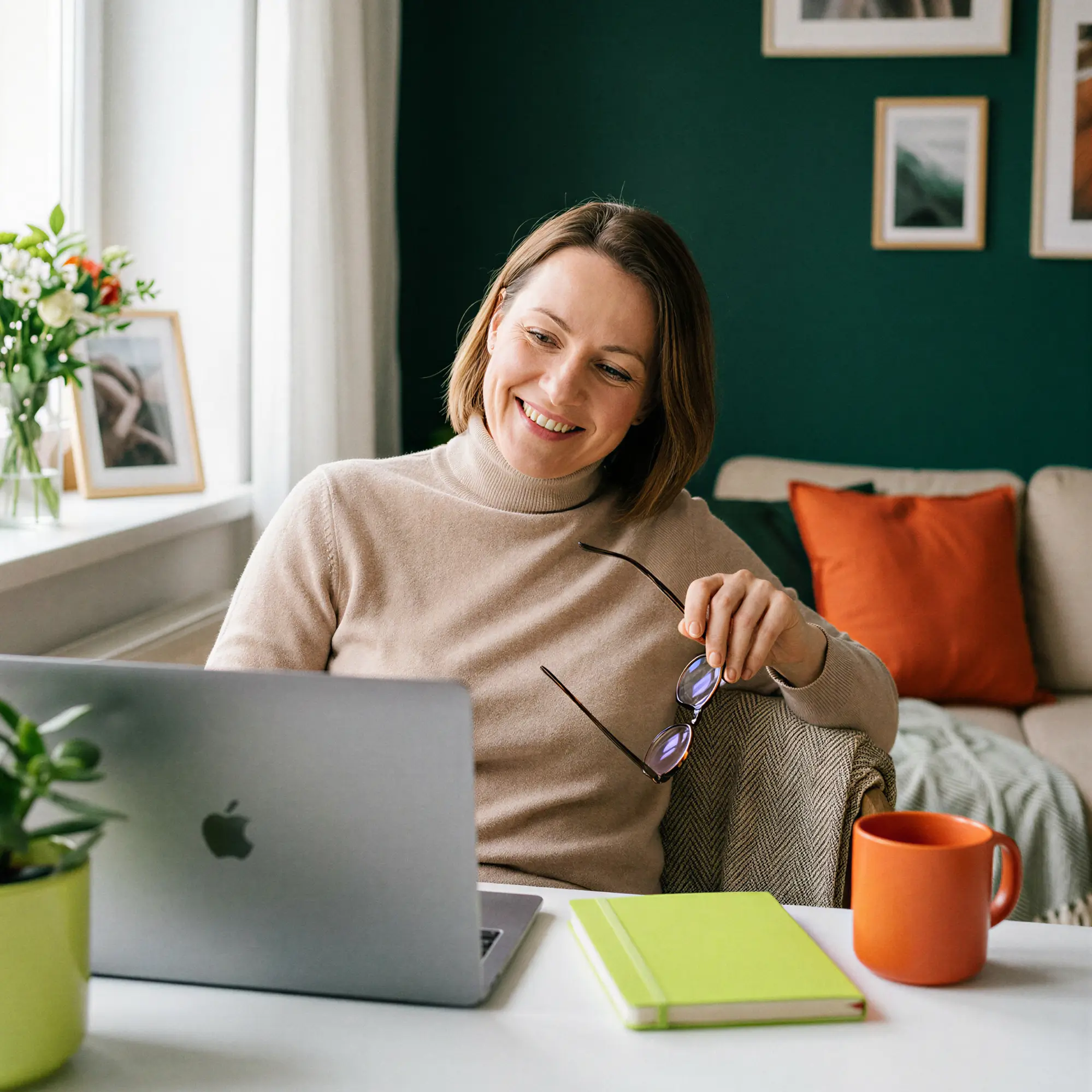 Woman smiling while holding glasses and looking at a laptop in a cozy living room with plants and orange cushions.