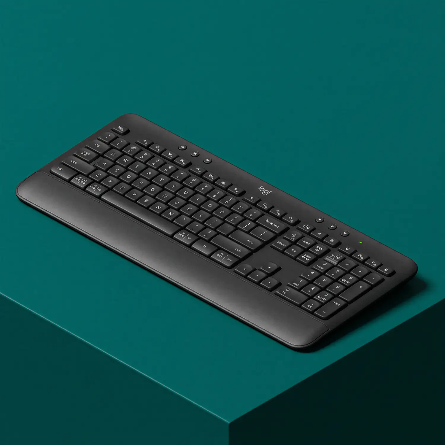 Black wireless computer keyboard with an integrated wrist rest on a teal surface.