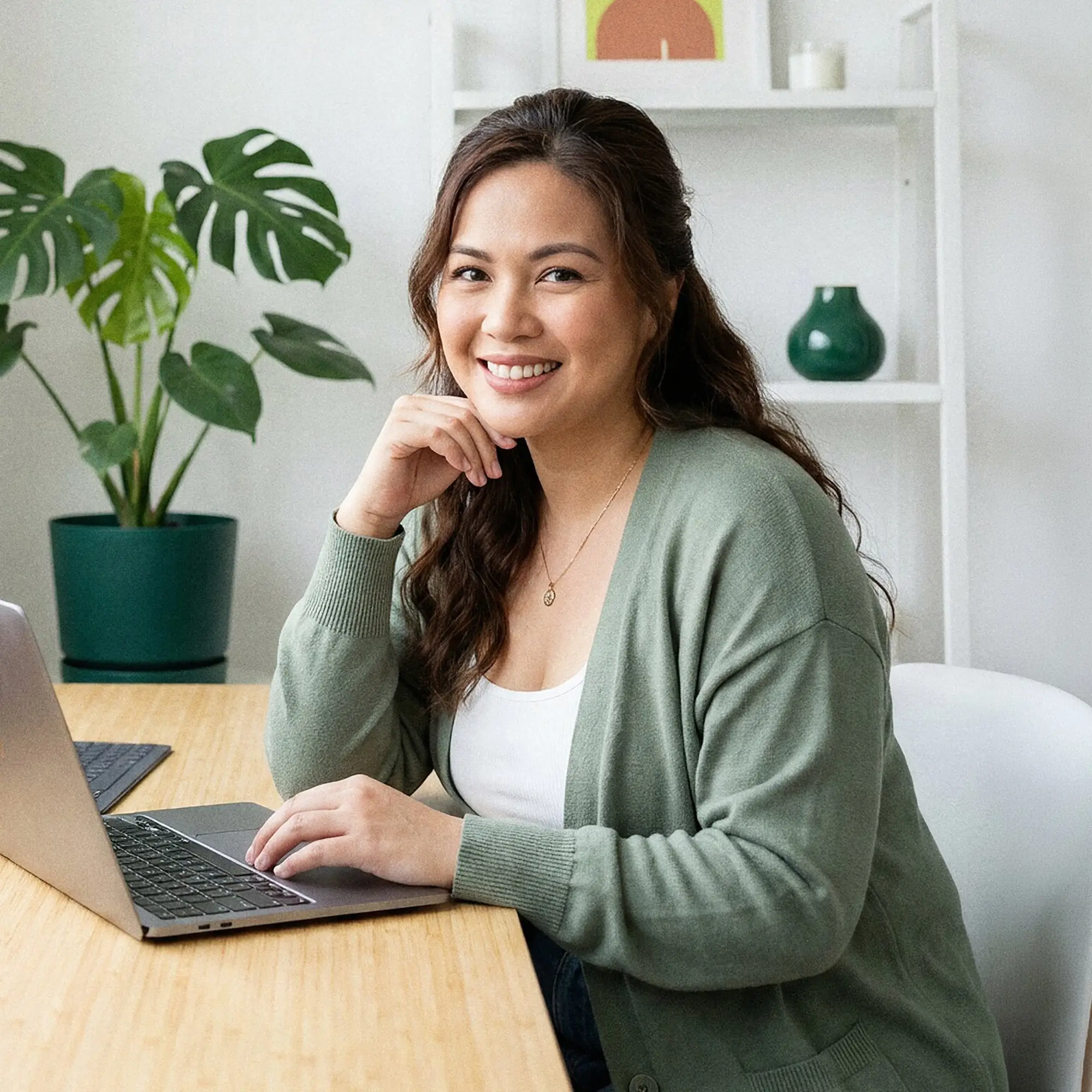 Smiling woman wearing a green cardigan working on a laptop at a wooden desk with a green potted plant and shelves in the background.